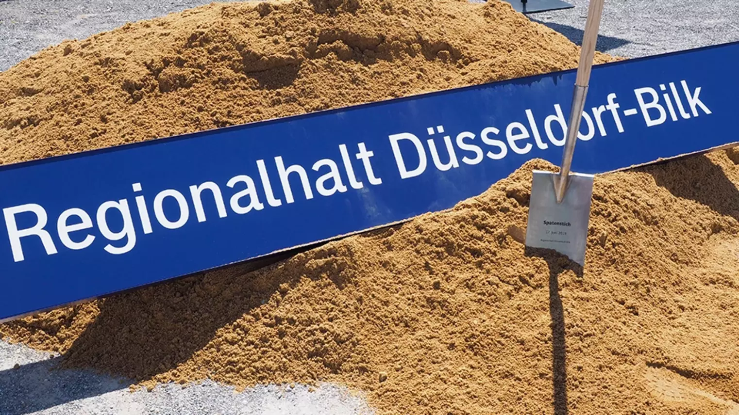 Das Bahnhofsschild von Düsseldorf-Bilk liegt im Sand, daneben ein Spaten