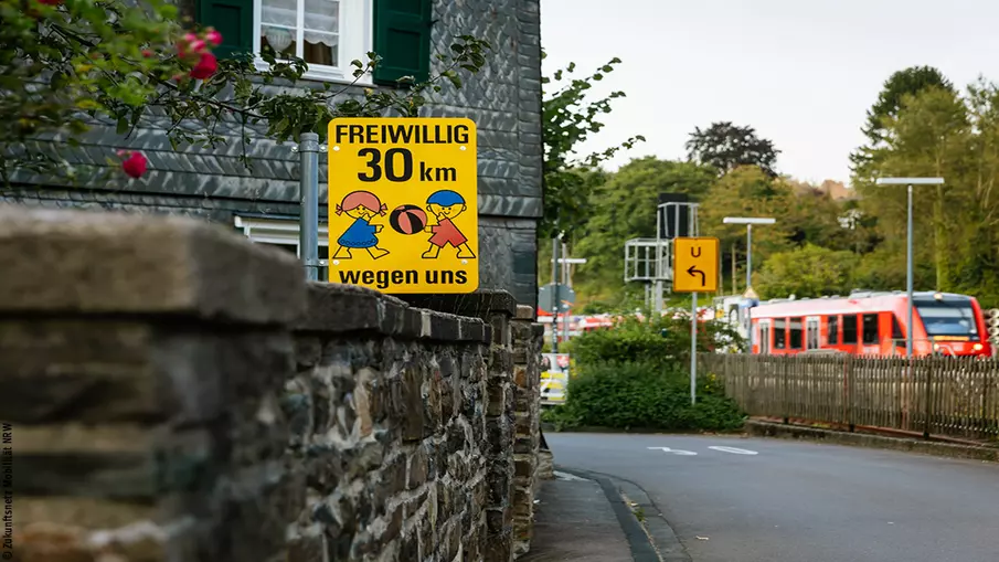 Blick auf eine Mauer, dahinter ein Haus, beides an einer 30-Zone. Im Hintergrund ist ein Regionalzug zu sehen.