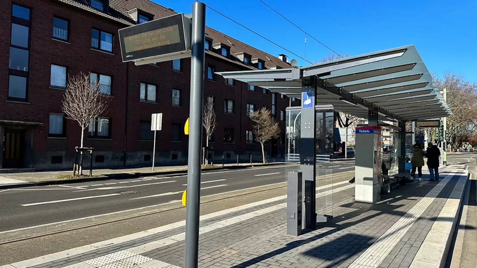 Neubau Haltestelle Brückelstraße: Blick auf die fertige Haltestelle mit Wetterschutz, dynamischen Fahrgastinformation, Ticketautomat und Sitzgelegenheiten, im Hintergrund sind zwei Fahrgäste zu sehen