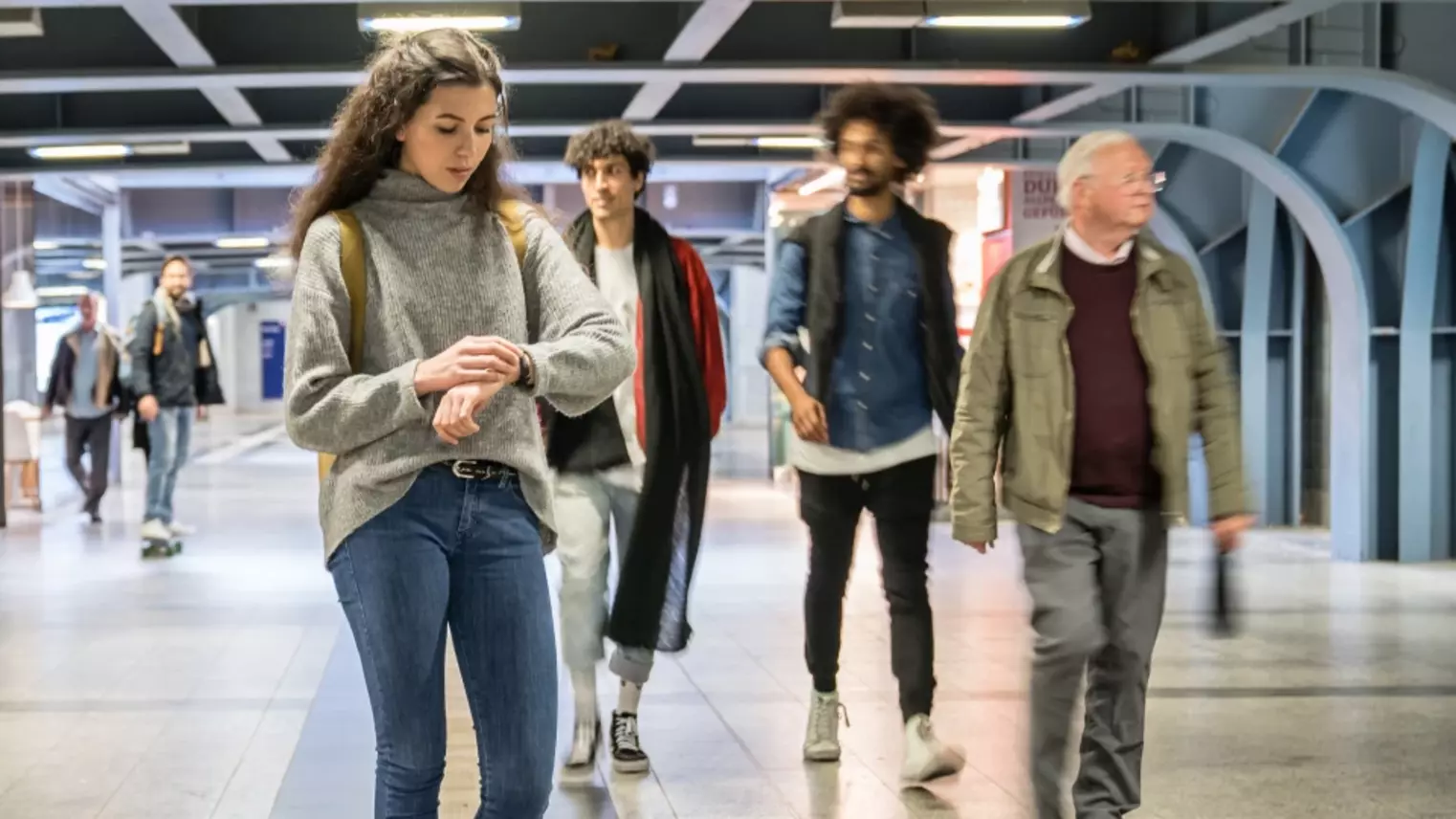 Personen in einem Bahnhof. Eine Frau im Vordergrund schaut auf ihre Armbanduhr