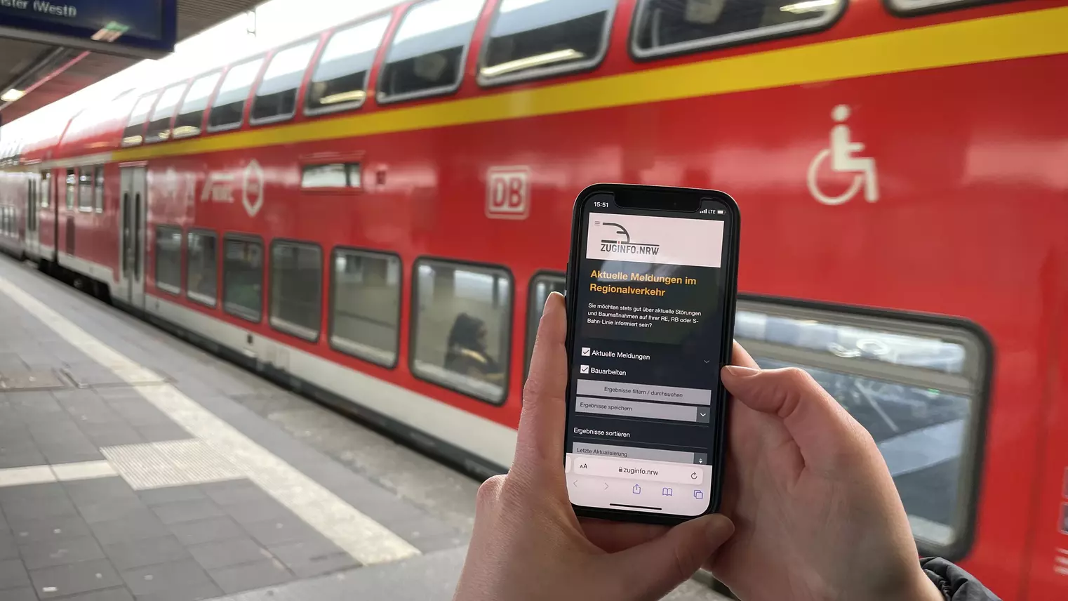 Ein Handy mit der Website zuginfo.nrw, im Hintergrund ein roter Zug