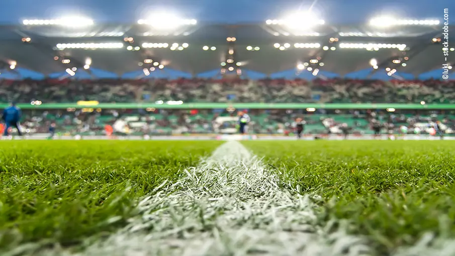 Der Fußballrasen und die Mittellinie stehen im Fokus des Bildes. Im Hintergrund ist eine Zuschauertribüne zu erkennen.