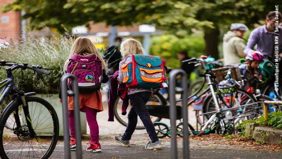 Zwei Schülerinnen mit Tornister laufen über einen Gehweg, im Hintergrund und Vordergrund sind abgestellte Fahrräder zu sehen