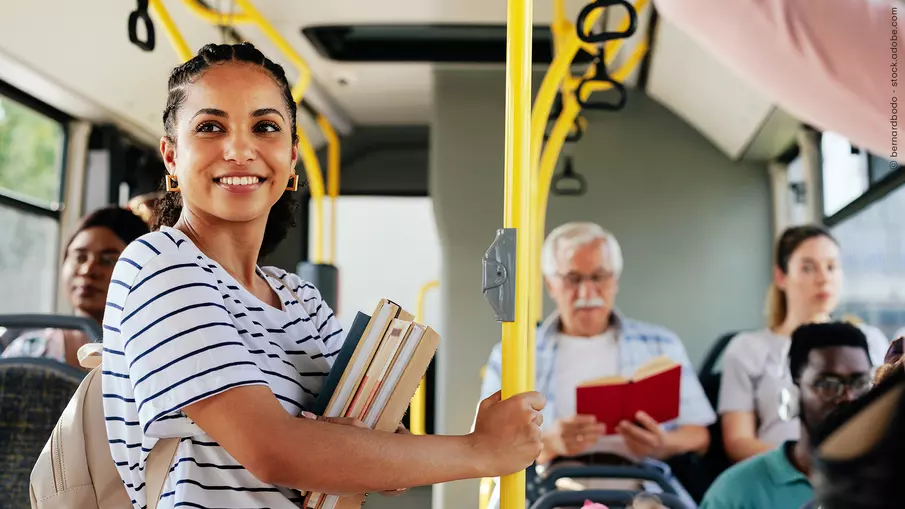 Eine junge Frau steht mit Büchern im Arm und Rucksack in einem Bus. Um sie herum weitere alte und junge Fahrgäste