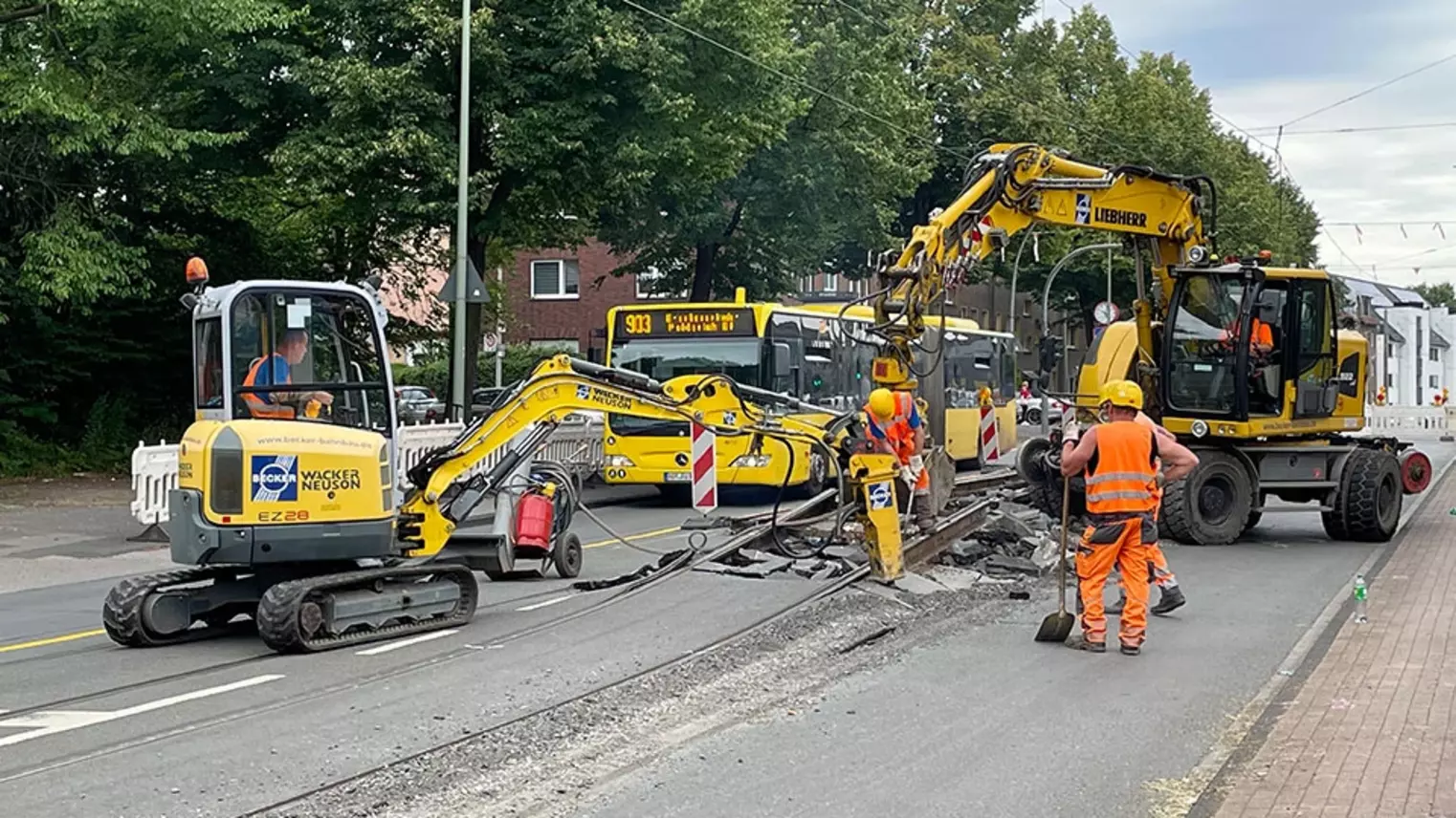 Neubau Haltestelle Brückelstraße: zwei Baumaschinen brechen den Asphalt auf und heben alte Gleise an, Bauarbeiter sind zu sehen, im Hintergrund ein Bus der DVG