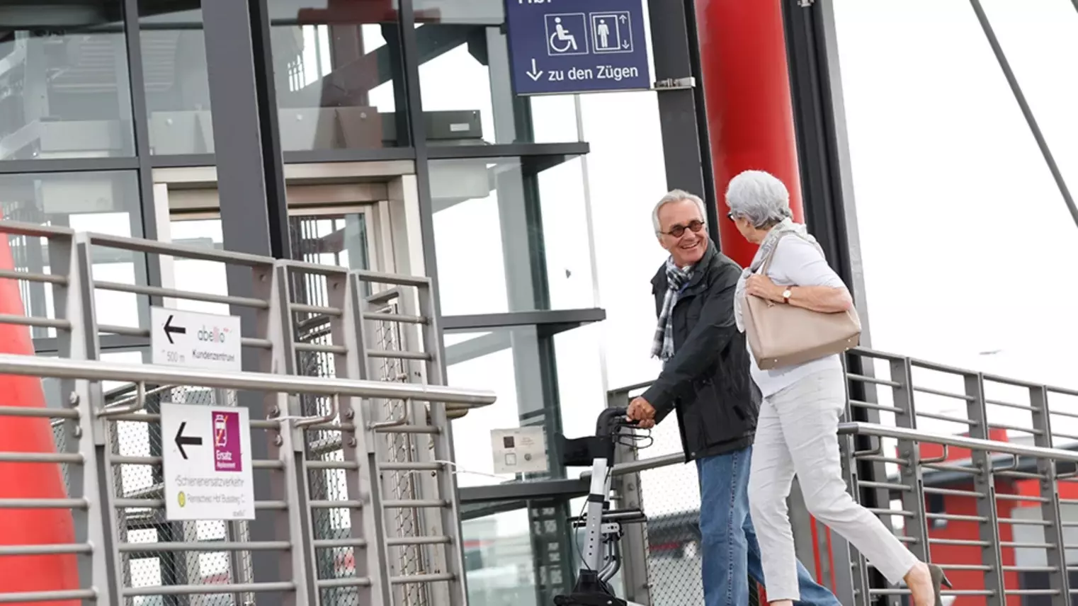 Ein älterer Fahrgast mit Rollator und seine weibliche Begleitung gehen am Bahnhof auf einen Aufzug zu