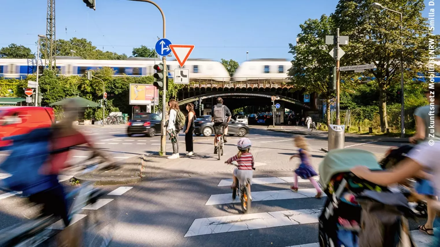 Blick auf eine Kreuzung: Kinder und Erwachsene mit Kinderwagen gehen über einen Zenbrastreifen, Fahrradfahrer auf einem Fahrradweg, Autos fahren über die Kreuzung, im Hintergrund ist ein Zug zu sehen