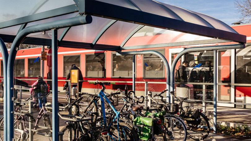 An einem Bahnsteig steht ein roter Zug, Fahrgäste steigen ein, im Vordergrund stehen Fahrräder unter einem Dach