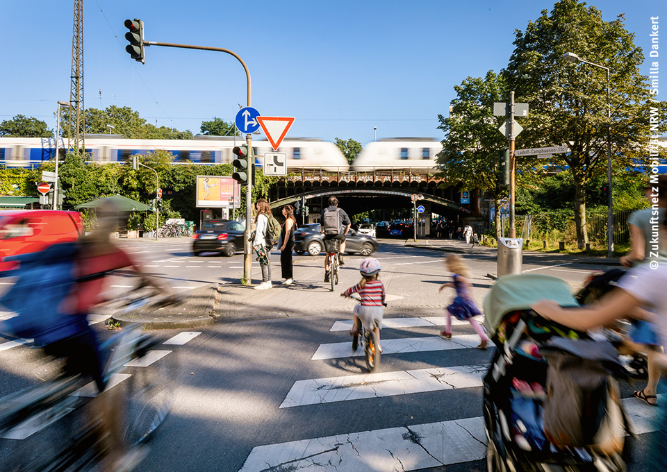 Blick auf eine Kreuzung: Kinder und Erwachsene mit Kinderwagen gehen über einen Zenbrastreifen, Fahrradfahrer auf einem Fahrradweg, Autos fahren über die Kreuzung, im Hintergrund ist ein Zug zu sehen